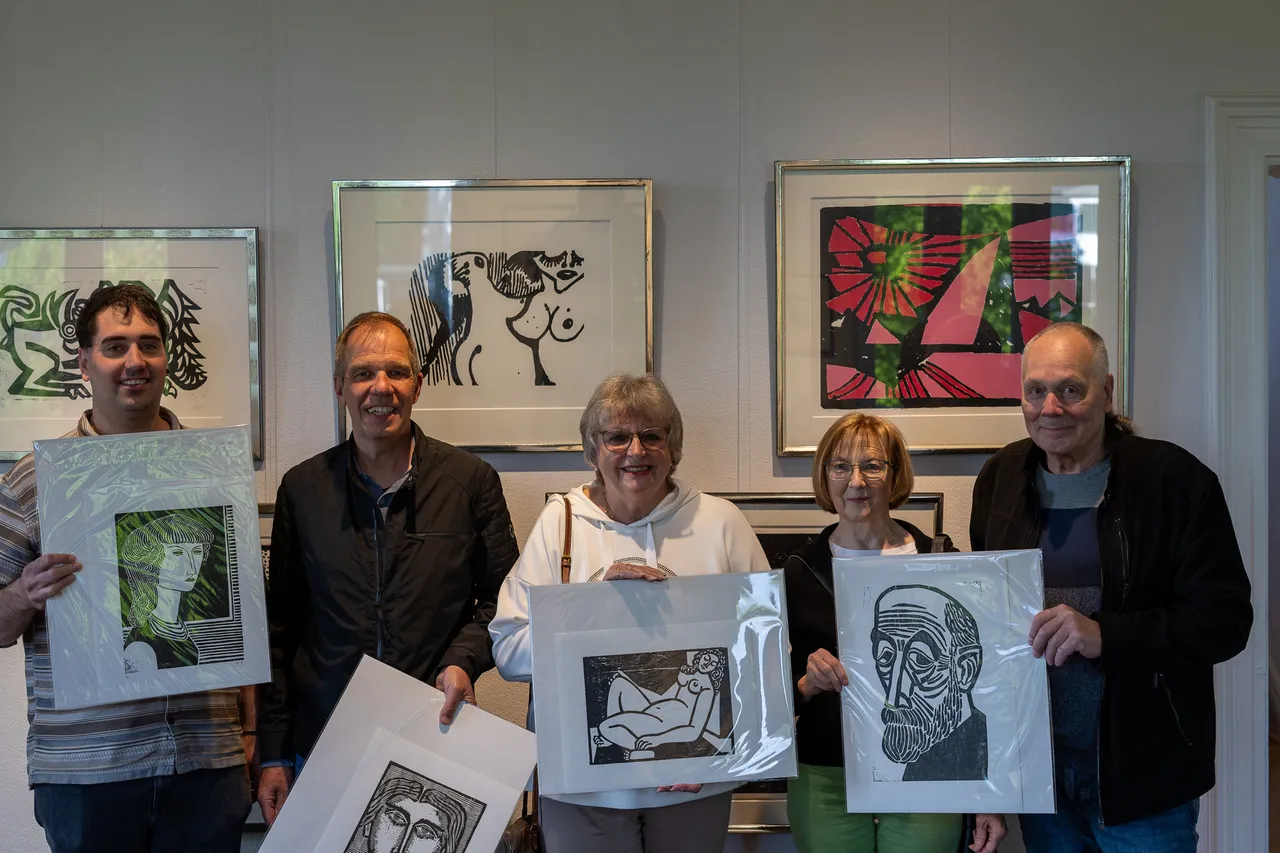 Fünf Personen halten jeweils ein gerahmtes Kunstwerk vor sich. Im Hintergrund hängen weitere Bilder an der Wand.