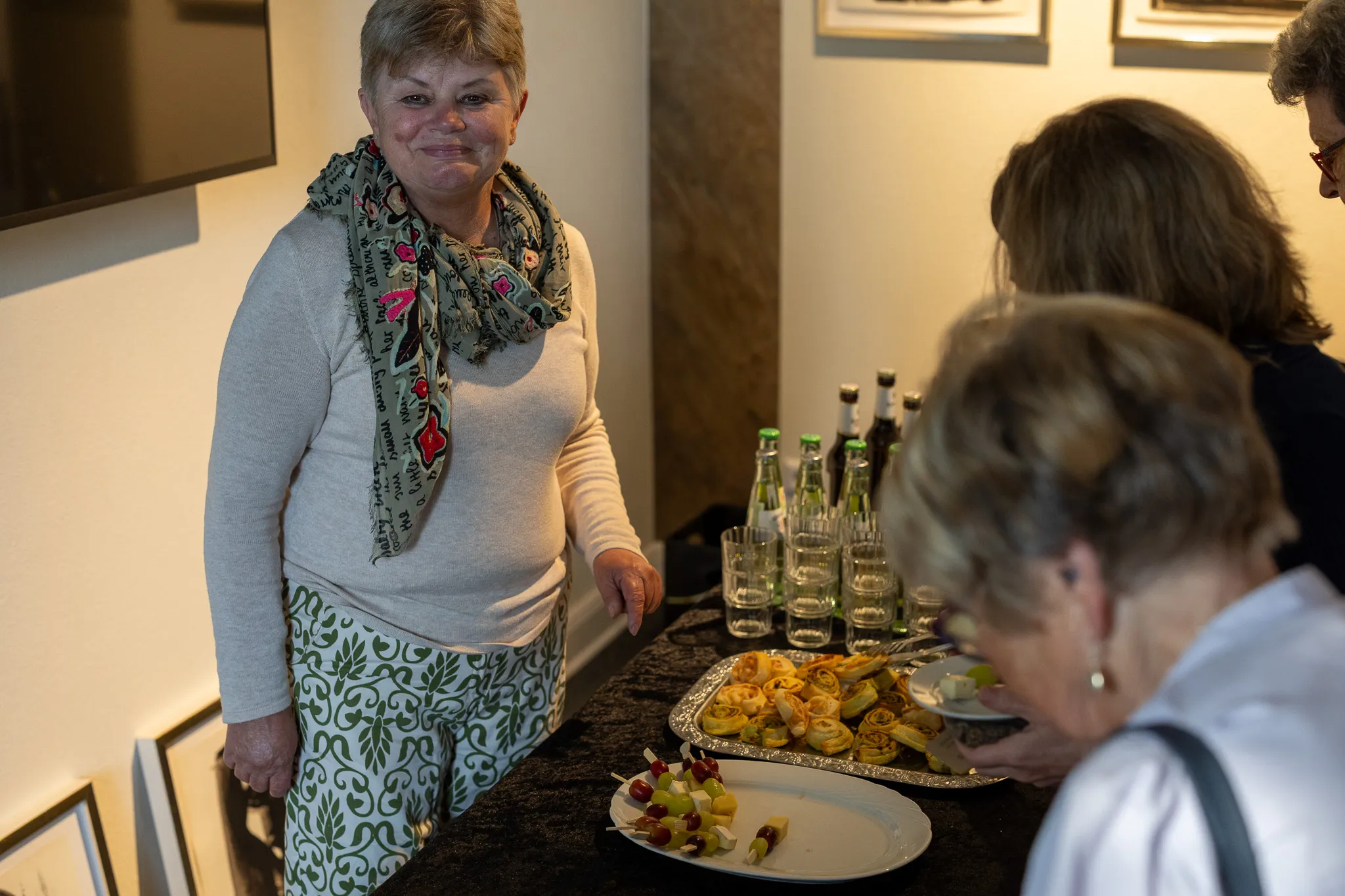 Eine Frau mit kurzen grauen Haaren lächelt in einem Raum, während andere Gäste im Hintergrund stehen und sich an einem Tisch mit Snacks bedienen.