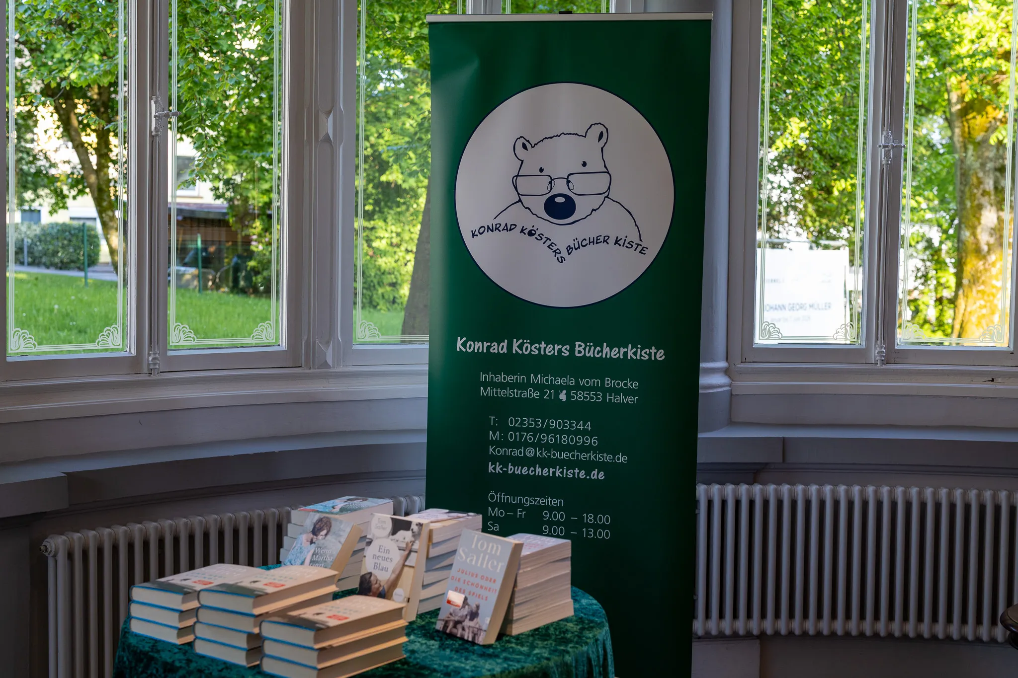 Schild der Buchhandlung Konrad Kösters Bücherkiste mit Bärenlogo und Kontaktinformationen vor Bücherstapel im Schaufenster.