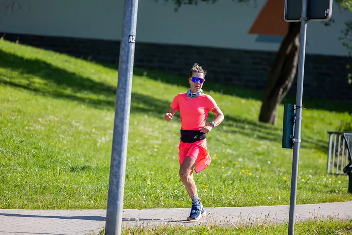Eine Frau mit hochgebundenem Haar und Sonnenbrille läuft auf einem Gehweg, gekleidet in leuchtend rosa Sportkleidung. Im Hintergrund eine Grünfläche und ein Laternenmast.