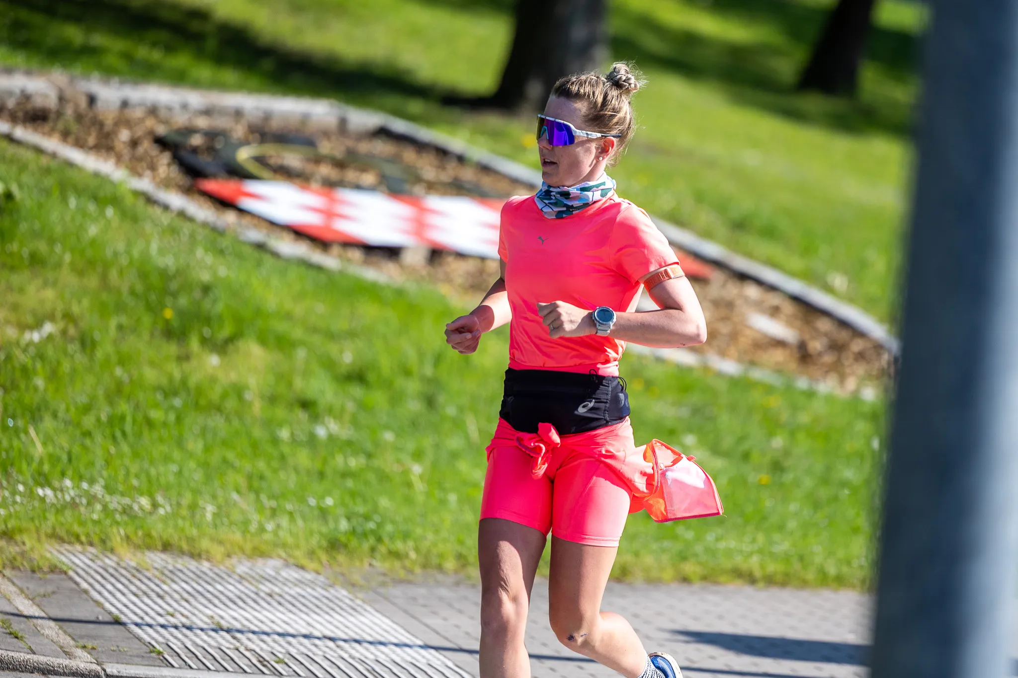 Eine Frau in pinker Sportkleidung läuft auf einem Gehweg, trägt eine Sonnenbrille und eine Uhr am Handgelenk. Im Hintergrund ist eine grüne Wiese und ein Baum zu sehen.