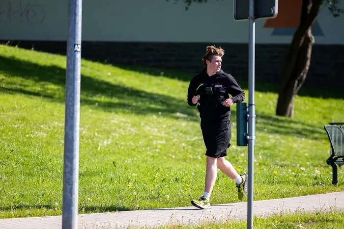 Ein junger Mann mit lockigem Haar joggt auf einem Gehweg neben einer Rasenfläche. Er trägt dunkle Sportkleidung und gelbe Laufschuhe.