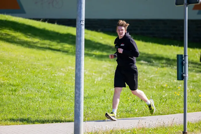 Ein junger Mann mit blonden Haaren joggt auf einem Gehweg neben einer grünen Wiese. Er trägt einen schwarzen Hoodie und kurze Hosen.