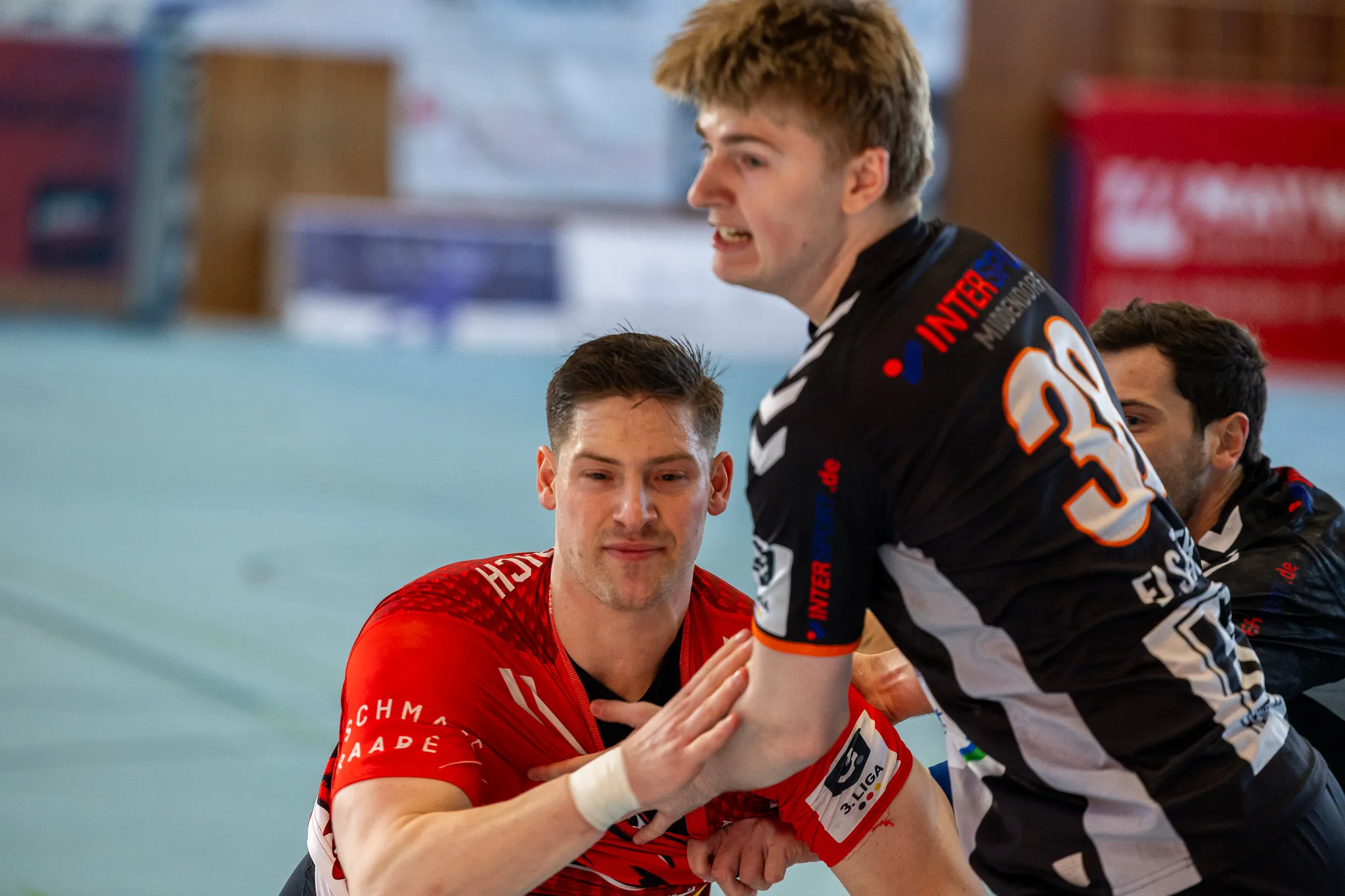 Zwei Handballspieler kämpfen um den Ball, während ein dritter im Hintergrund steht. Der Spieler im roten Trikot blickt entschlossen, der im schwarzen Trikot schreit. Im Hintergrund ist ein Werbebanner für die 3. Liga zu sehen.