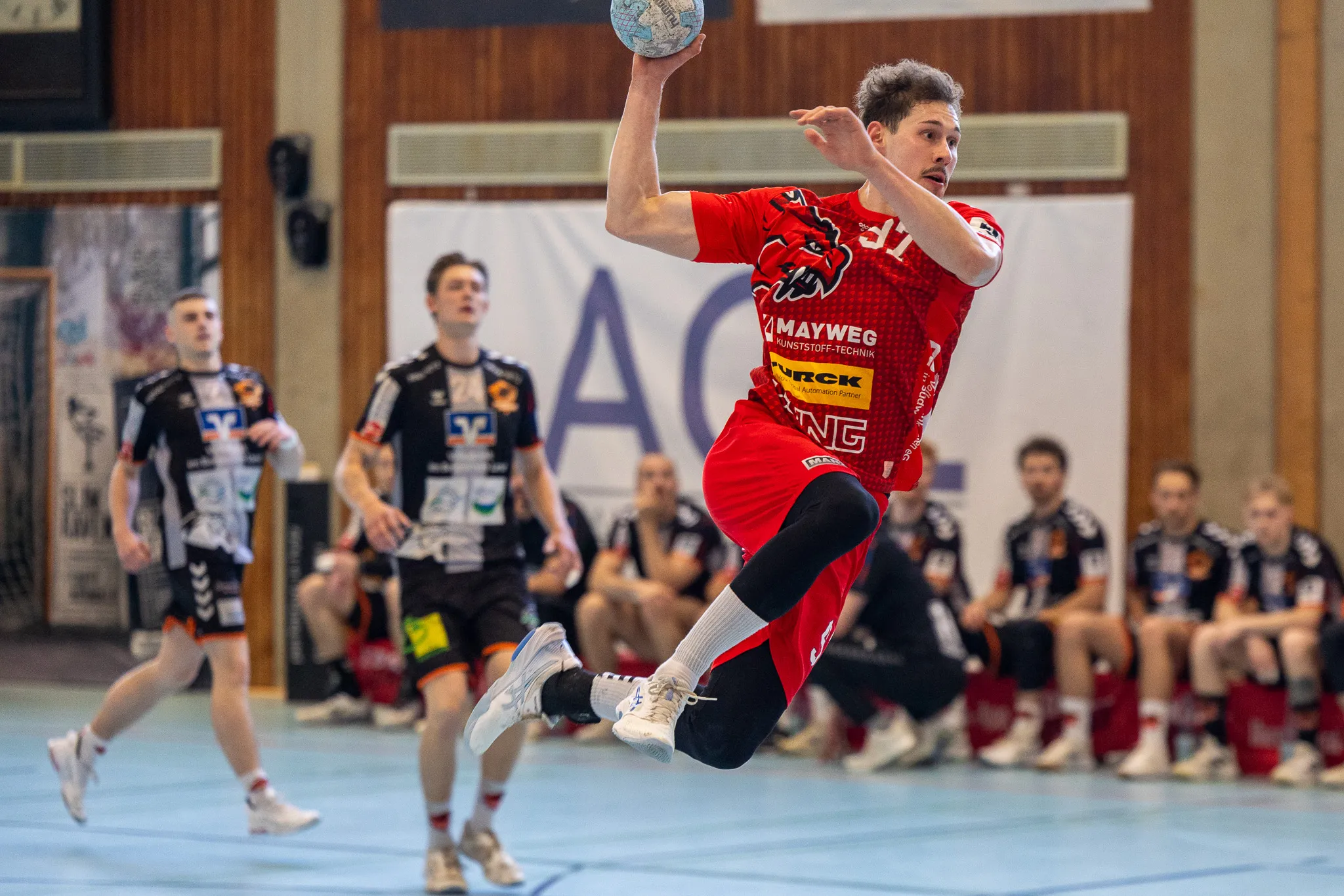 Handballspieler in rotem Trikot wirft den Ball während eines Spiels in einer Sporthalle. Im Hintergrund weitere Spieler und Zuschauer.