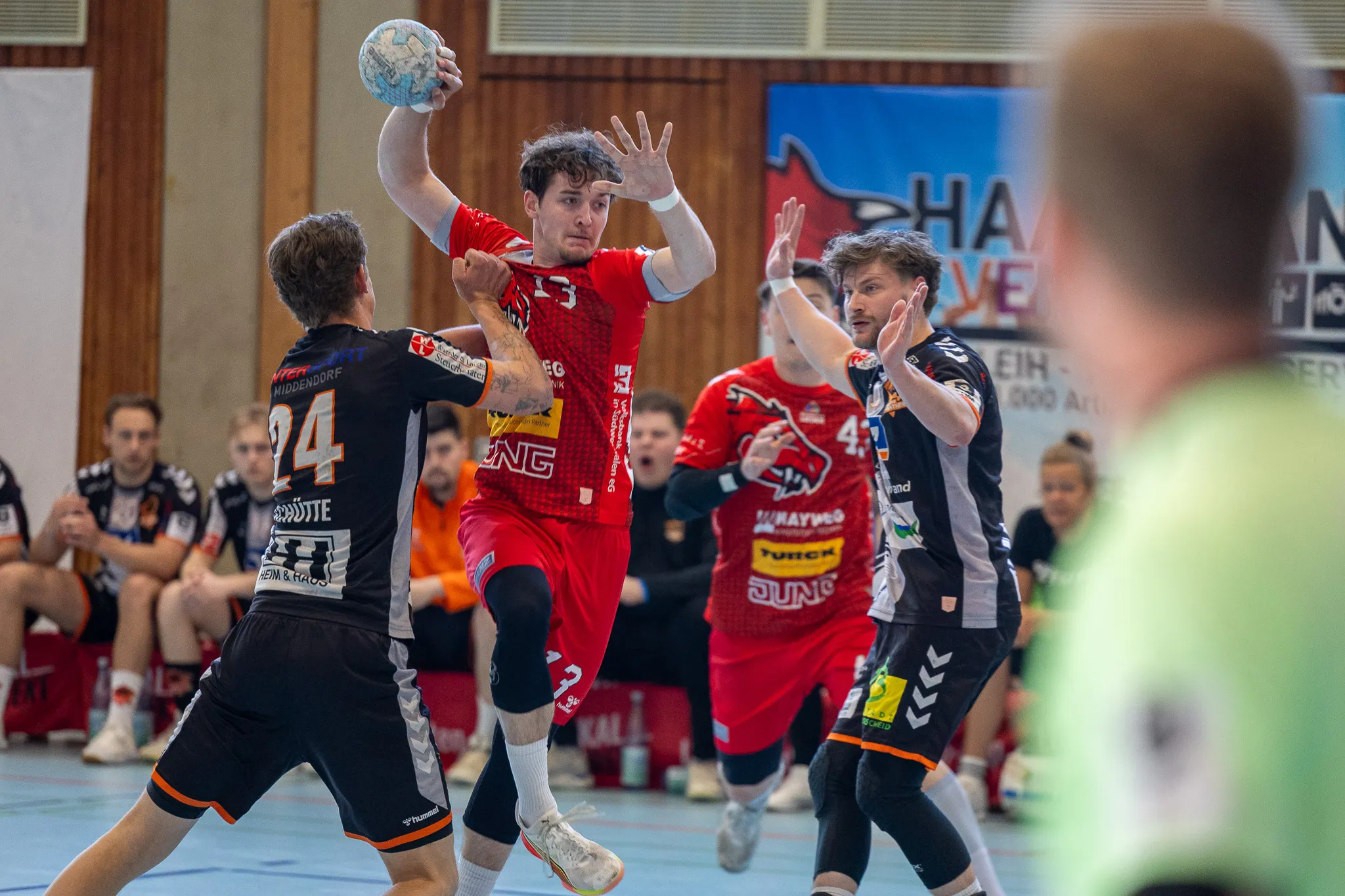 Handballspieler in Rot und Schwarz werfen und blocken den Ball während eines Spiels in einer Sporthalle.