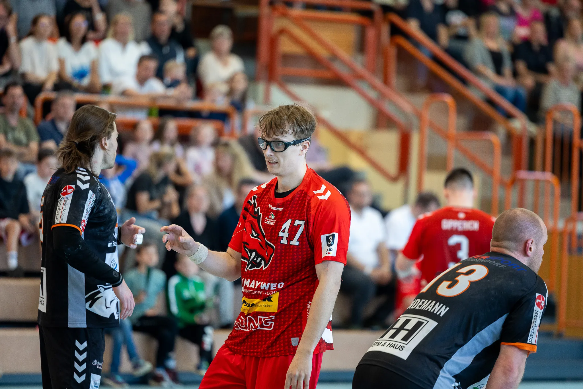 Zwei Handballspieler der SGSH Dragons und ein Spieler der Bergischen Panther stehen sich während eines Spiels gegenüber. Der Spieler der Dragons trägt eine Brille und das Trikot mit der Nummer 42.