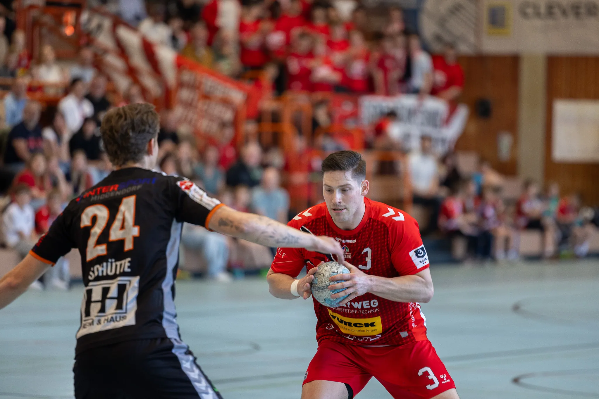 Handballspieler in roten Trikots mit der Nummer 3 wirft den Ball während eines Spiels gegen einen Gegner mit der Nummer 24 im schwarzen und orangefarbenen Trikot.