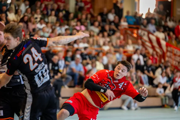Handballspieler in roten Trikots mit der Nummer 4 und 24 im Spiel, umgeben von Zuschauern in einer Sporthalle.