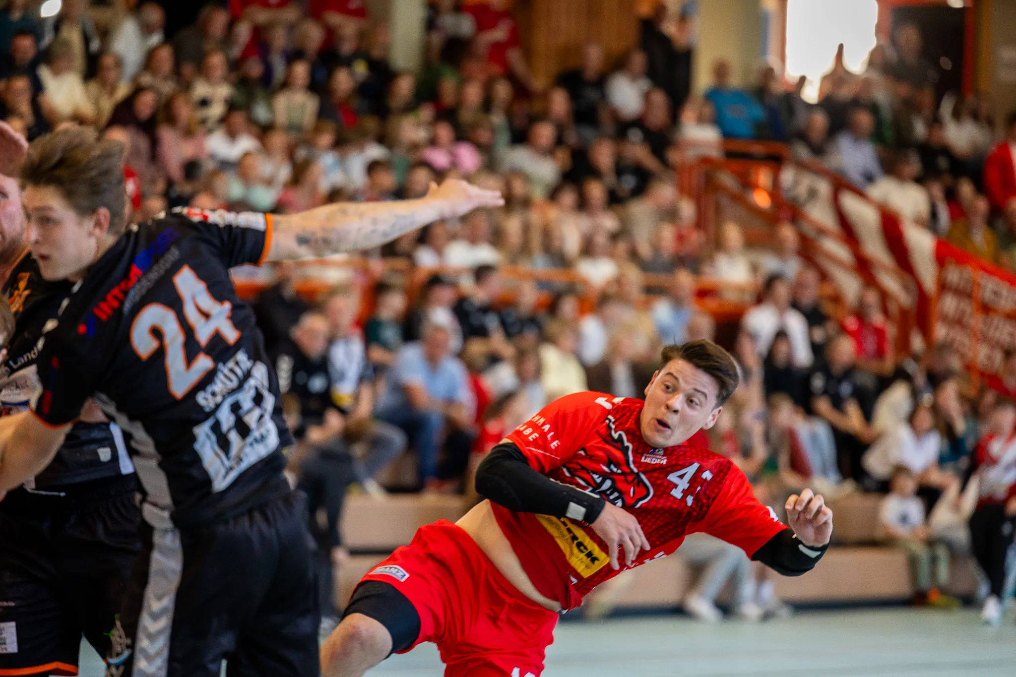 Handballspieler in roten Trikots mit der Nummer 4 und 24 im Spiel, umgeben von Zuschauern in einer Sporthalle.