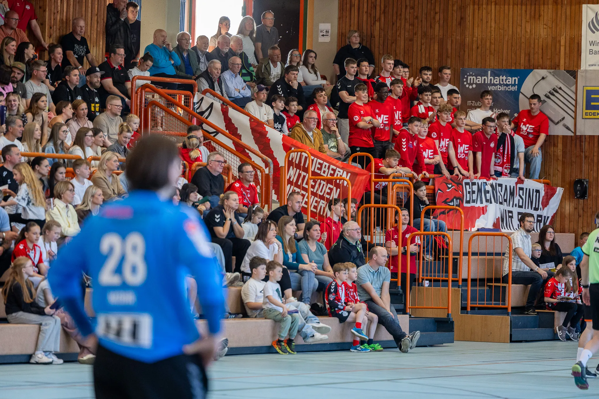 Zuschauer in einem rot-schwarzen Farbschema feuern ein Handballspiel an, während ein Spieler mit der Trikotnummer 28 im Vordergrund steht.