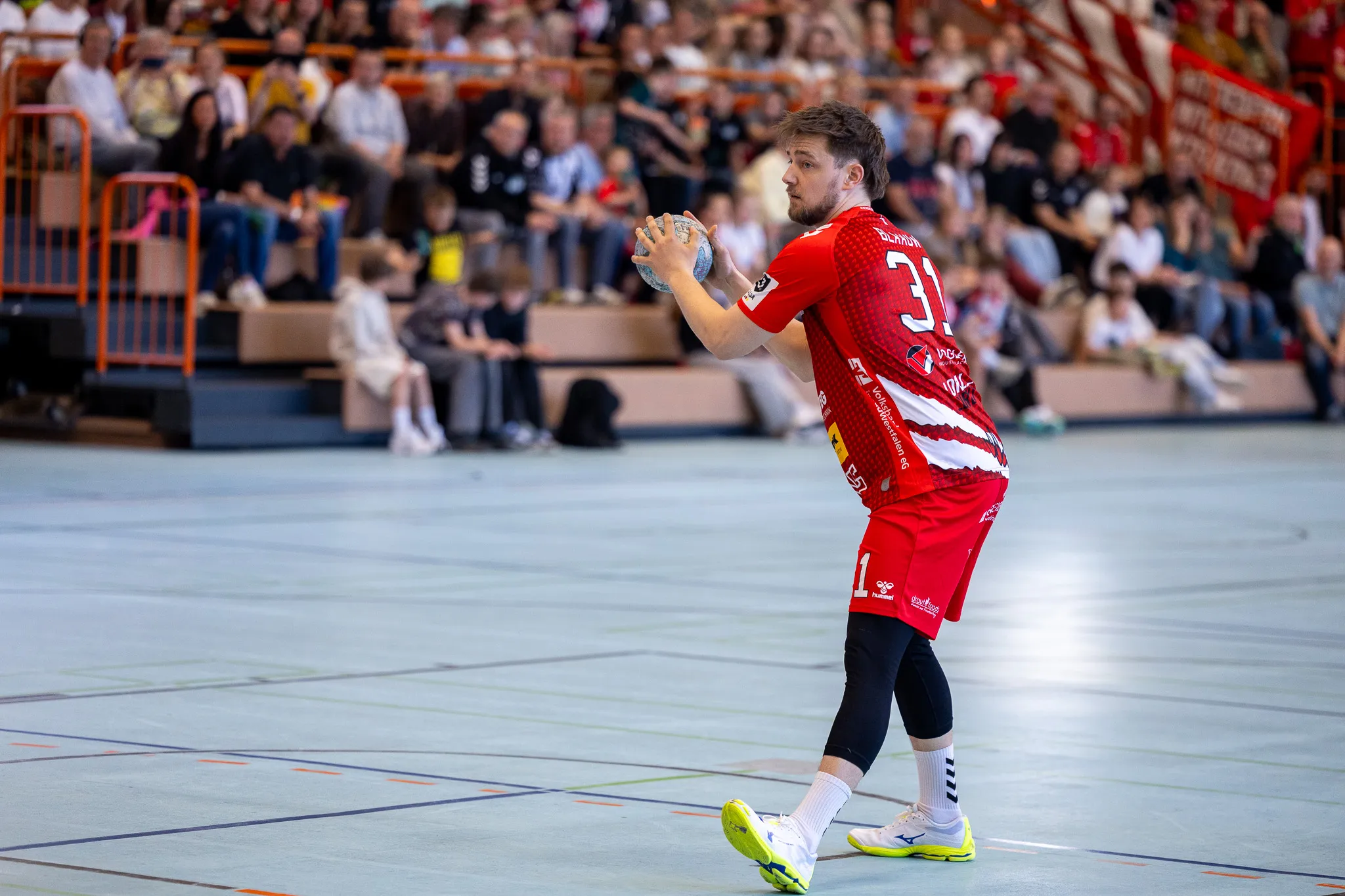 Handballspieler in rotem Trikot mit der Nummer 31 wirft den Ball während eines Spiels in einer vollbesetzten Halle.