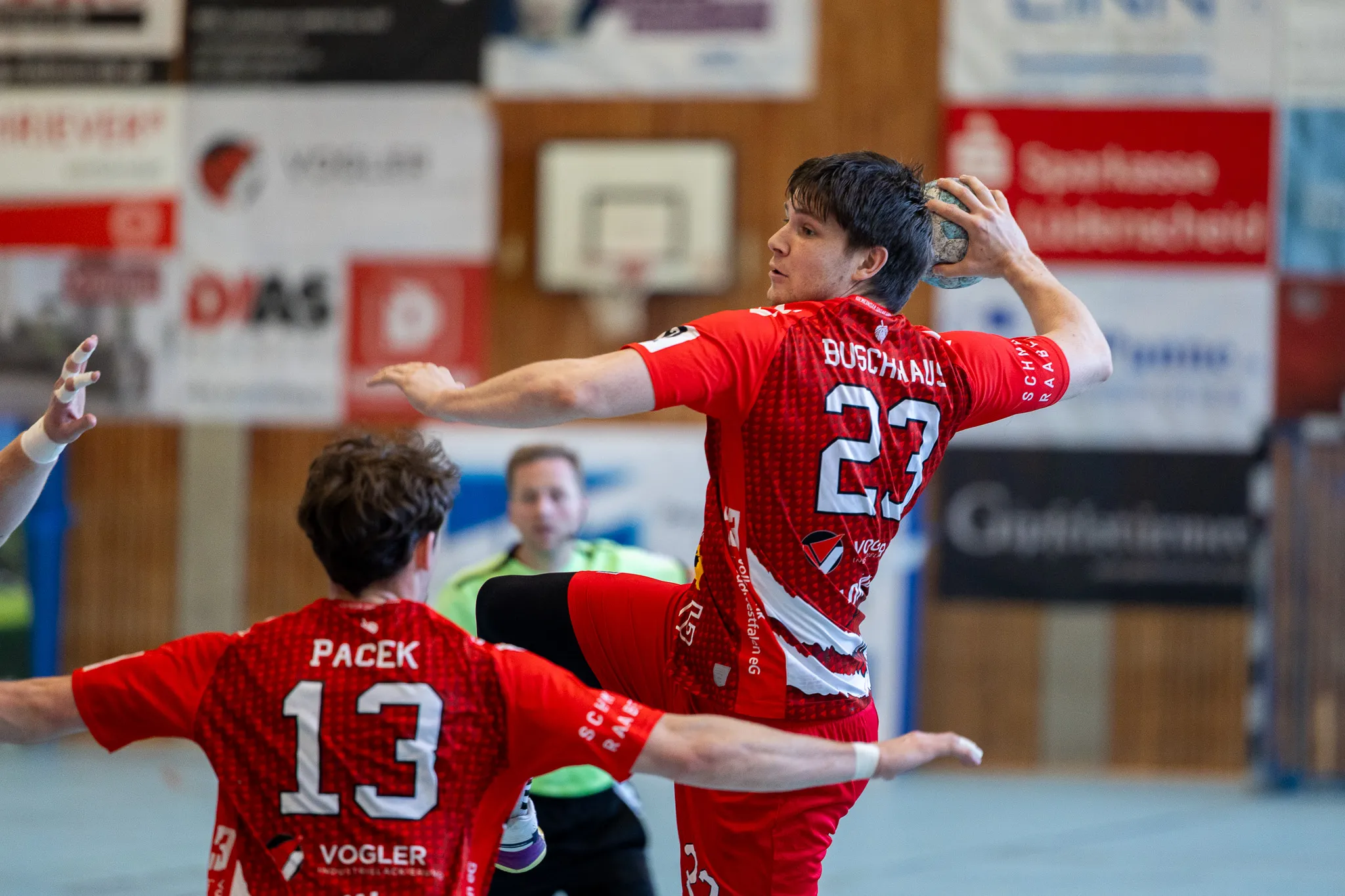 Handballspieler in roten Trikots mit den Nummern 23 und 13 in Aktion während eines Spiels in einer Sporthalle.