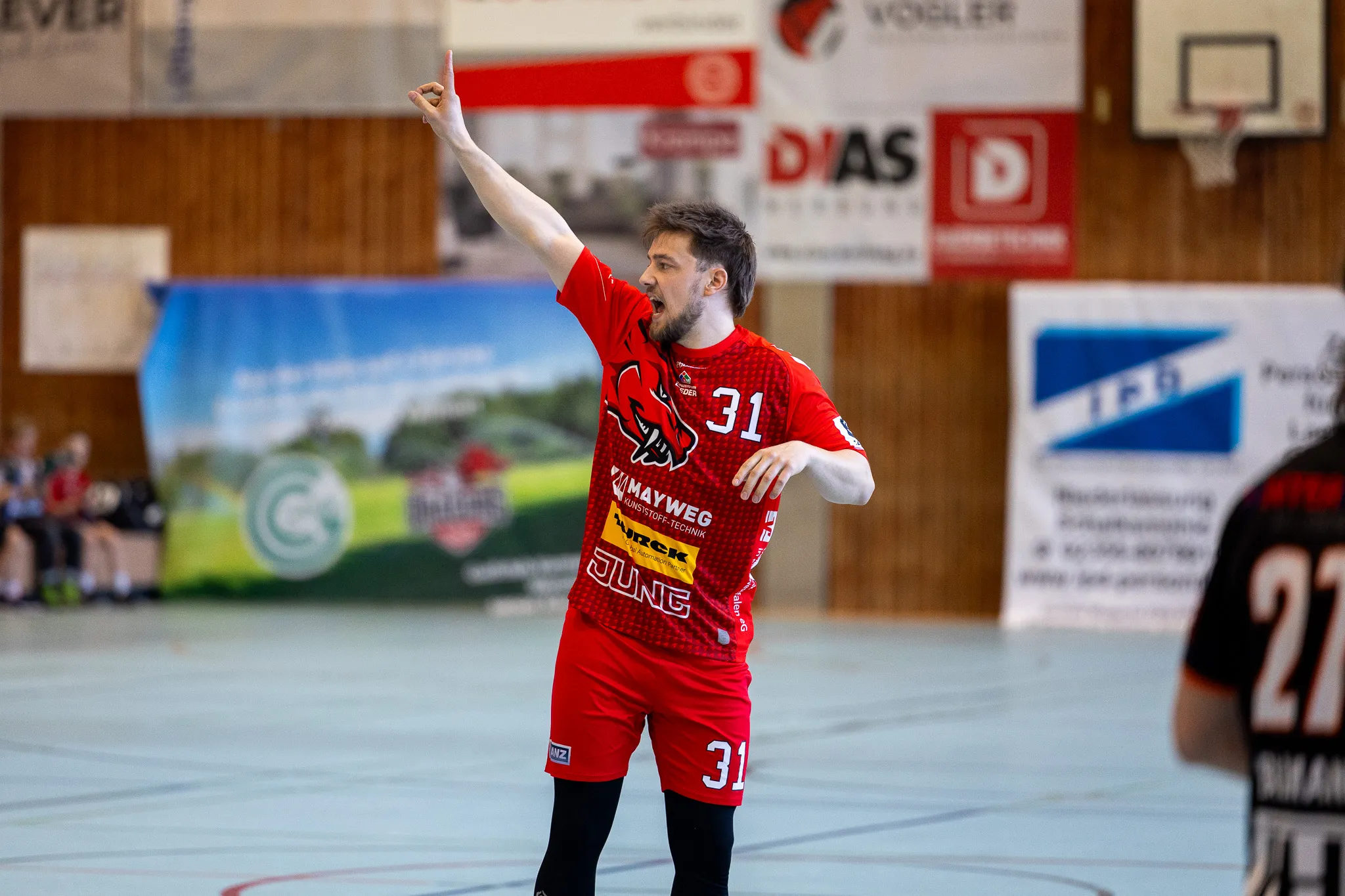 Handballspieler in rotem Trikot mit der Nummer 31 jubelt mit erhobener Faust in einer Sporthalle. Im Hintergrund sind weitere Spieler und Zuschauer zu sehen.