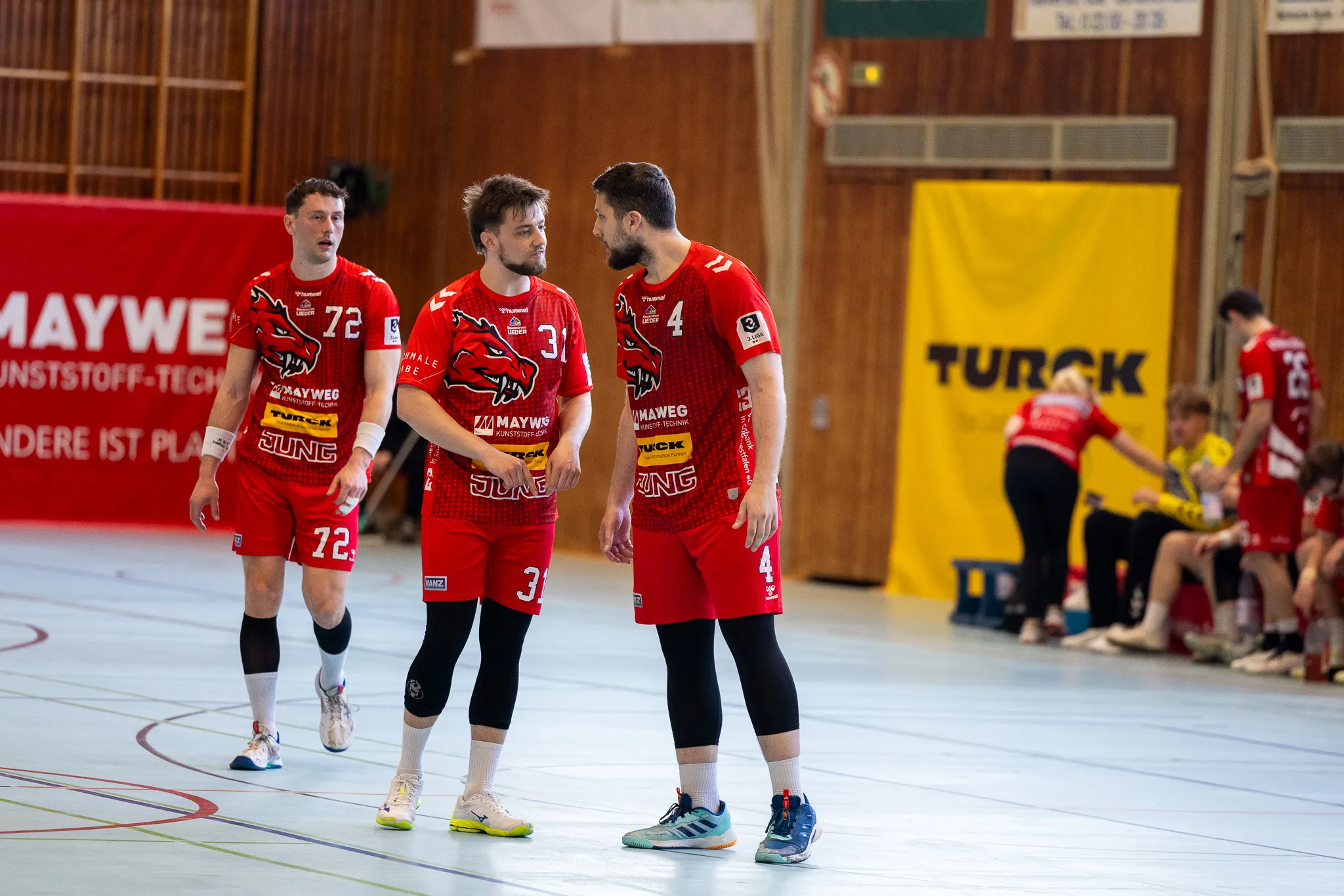 Drei Handballspieler der SGSH Dragons in roten Trikots stehen auf dem Spielfeld und besprechen etwas, während im Hintergrund weitere Spieler zu sehen sind. Im Hintergrund ein Werbebanner von Turck.