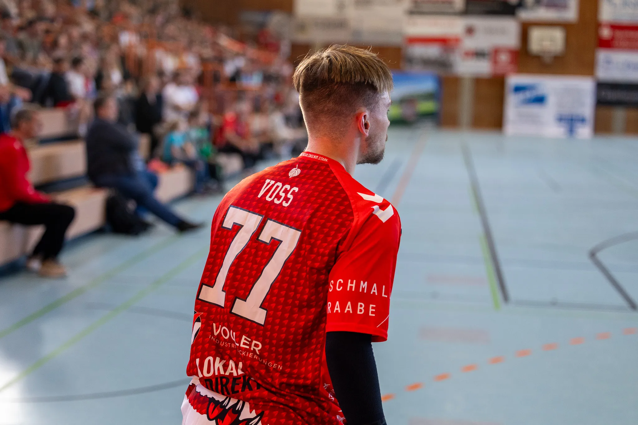 Handballspieler mit der Trikotnummer 77 geht auf dem Spielfeld, im Hintergrund weitere Spieler und Zuschauerränge.