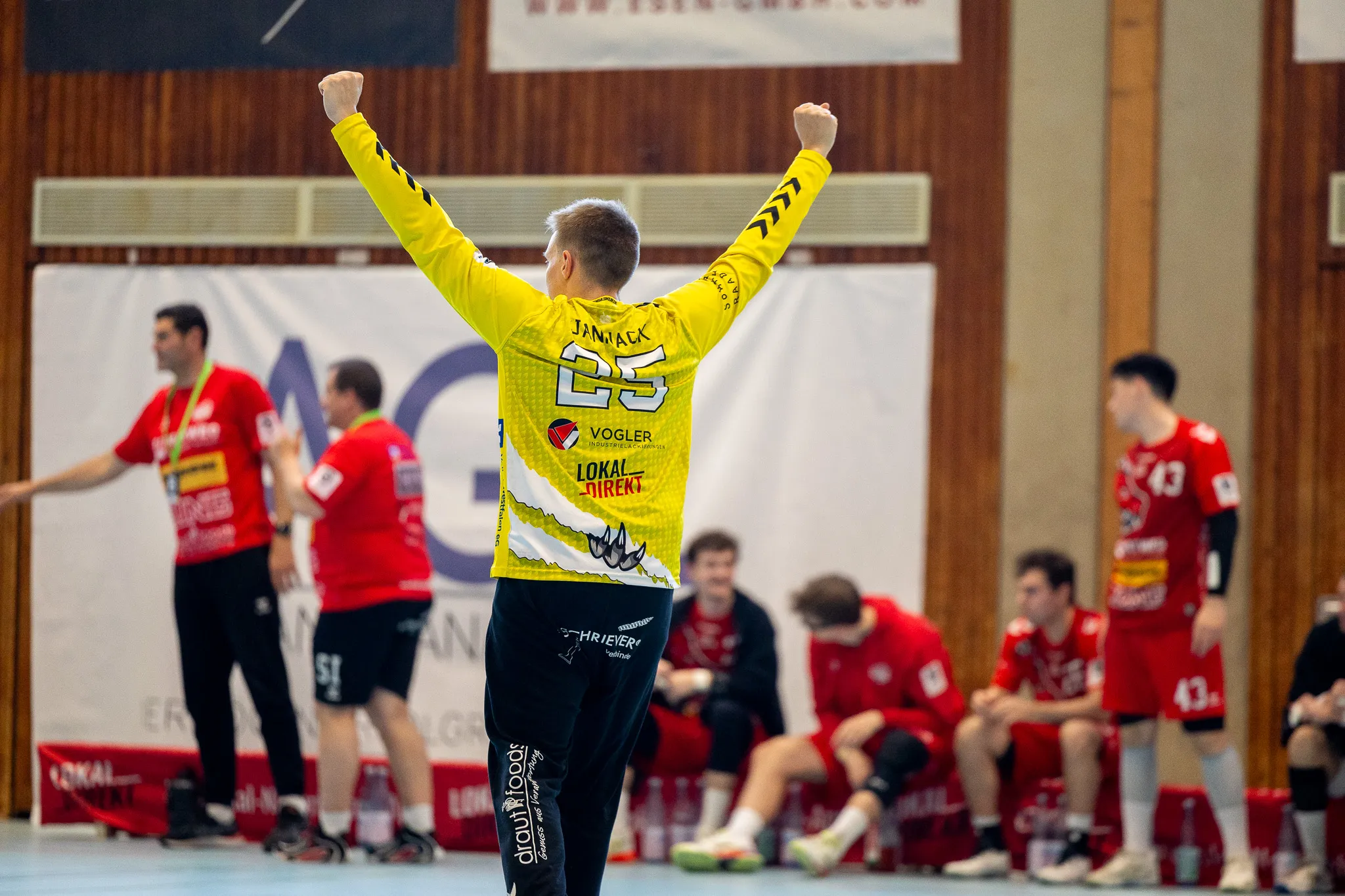 Handballspieler mit der Nummer 25 und dem Schriftzug „Jannack“ auf dem Rücken jubelt mit erhobenen Armen in einer Sporthalle. Im Hintergrund stehen weitere Spieler in roten Trikots.