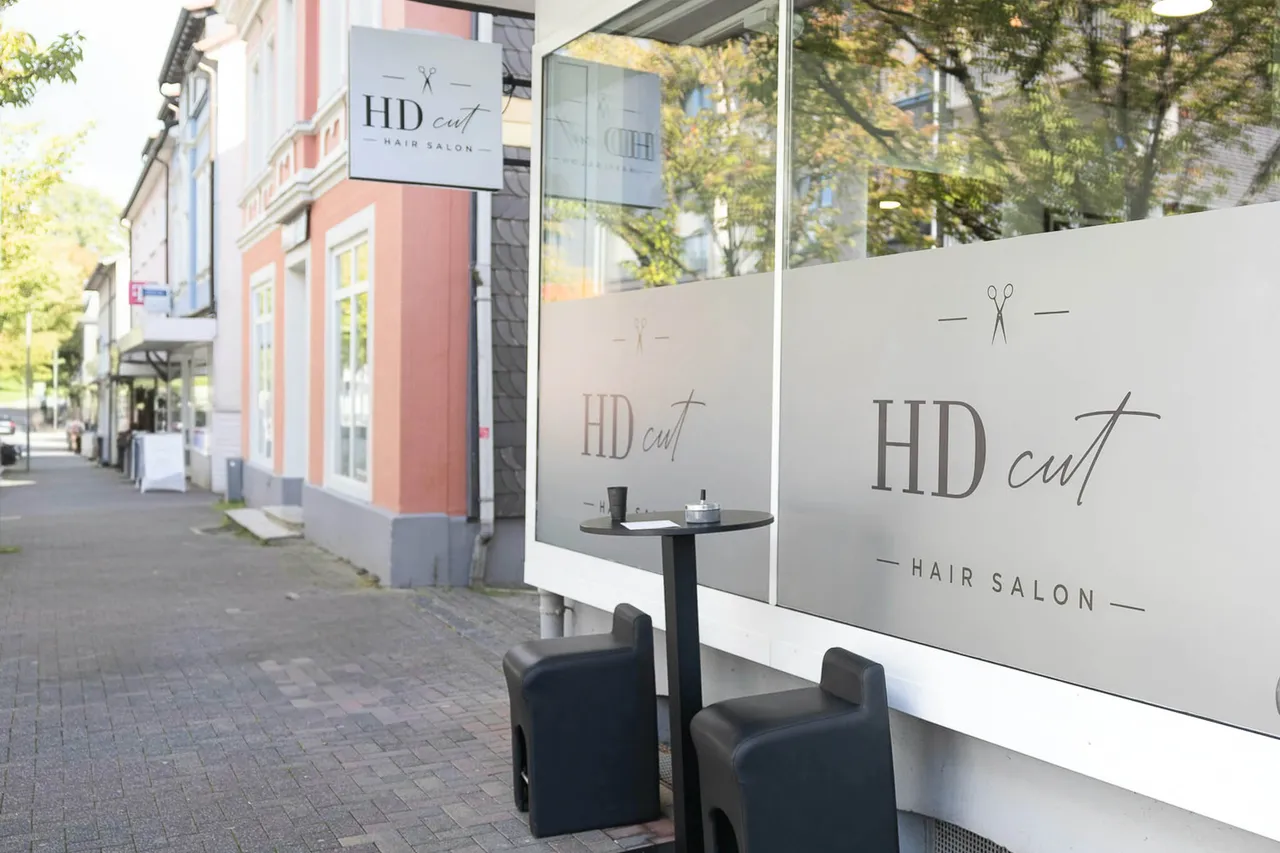 Außenansicht des Friseursalons „HDcut“ mit Logo auf der Fensterfront und einem kleinen Sitzbereich auf dem Gehweg.
