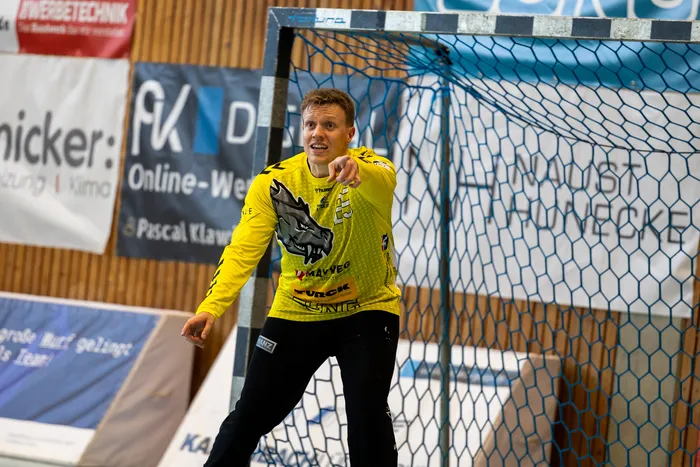Handballtorwart in gelbem Trikot mit Wolfskopfmotiv gestikuliert vor dem Tor, im Hintergrund Werbebanner und Zuschauerränge.