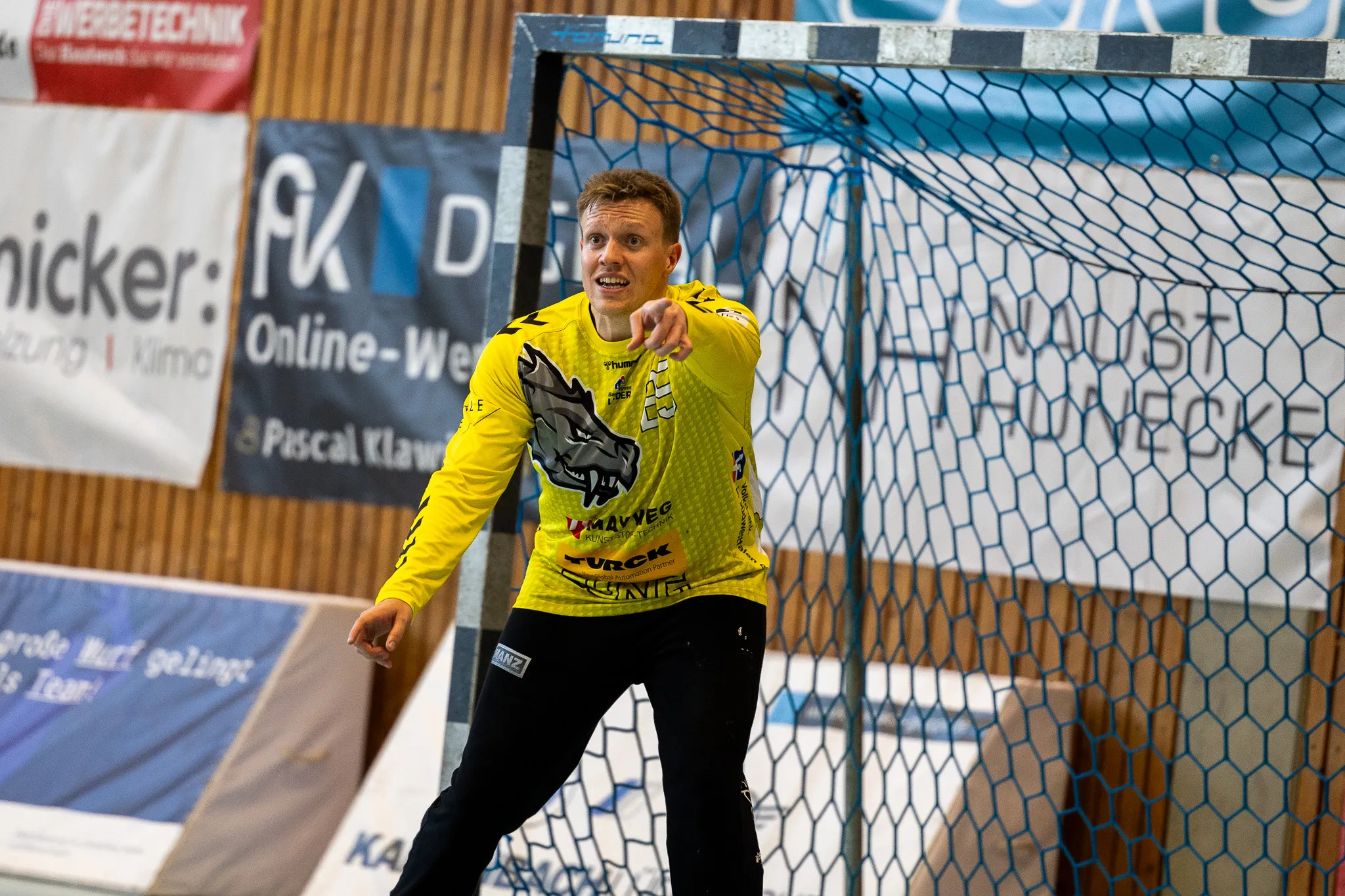 Handballtorwart in gelbem Trikot mit Wolfskopfmotiv gestikuliert vor dem Tor, im Hintergrund Werbebanner und Zuschauerränge.