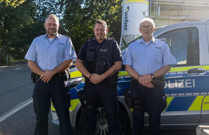 Drei Polizisten in Uniform stehen vor einem blau-gelben Streifenwagen.