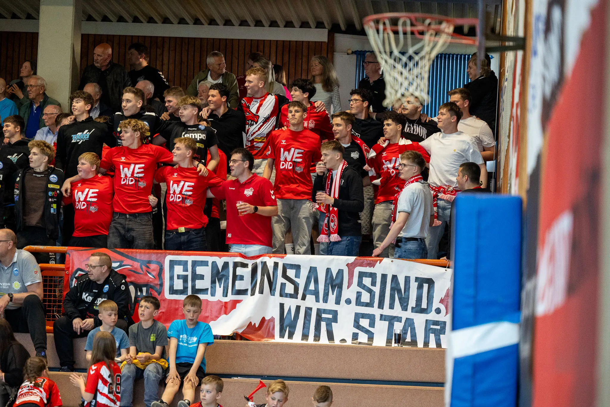 Zuschauer jubeln bei einem Handballspiel, viele tragen rote T-Shirts mit der Aufschrift „WE DID IT“. Ein Banner mit dem Slogan „Gemeinsam sind wir Star“ ist im Vordergrund zu sehen.
