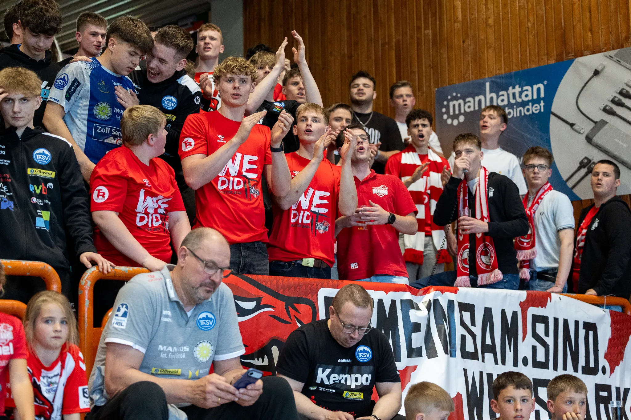 Jubelnde Fans in roten Trikots und Schals feiern in einer Sporthalle, im Hintergrund ein Banner mit der Aufschrift „MENSAM!SIND.“