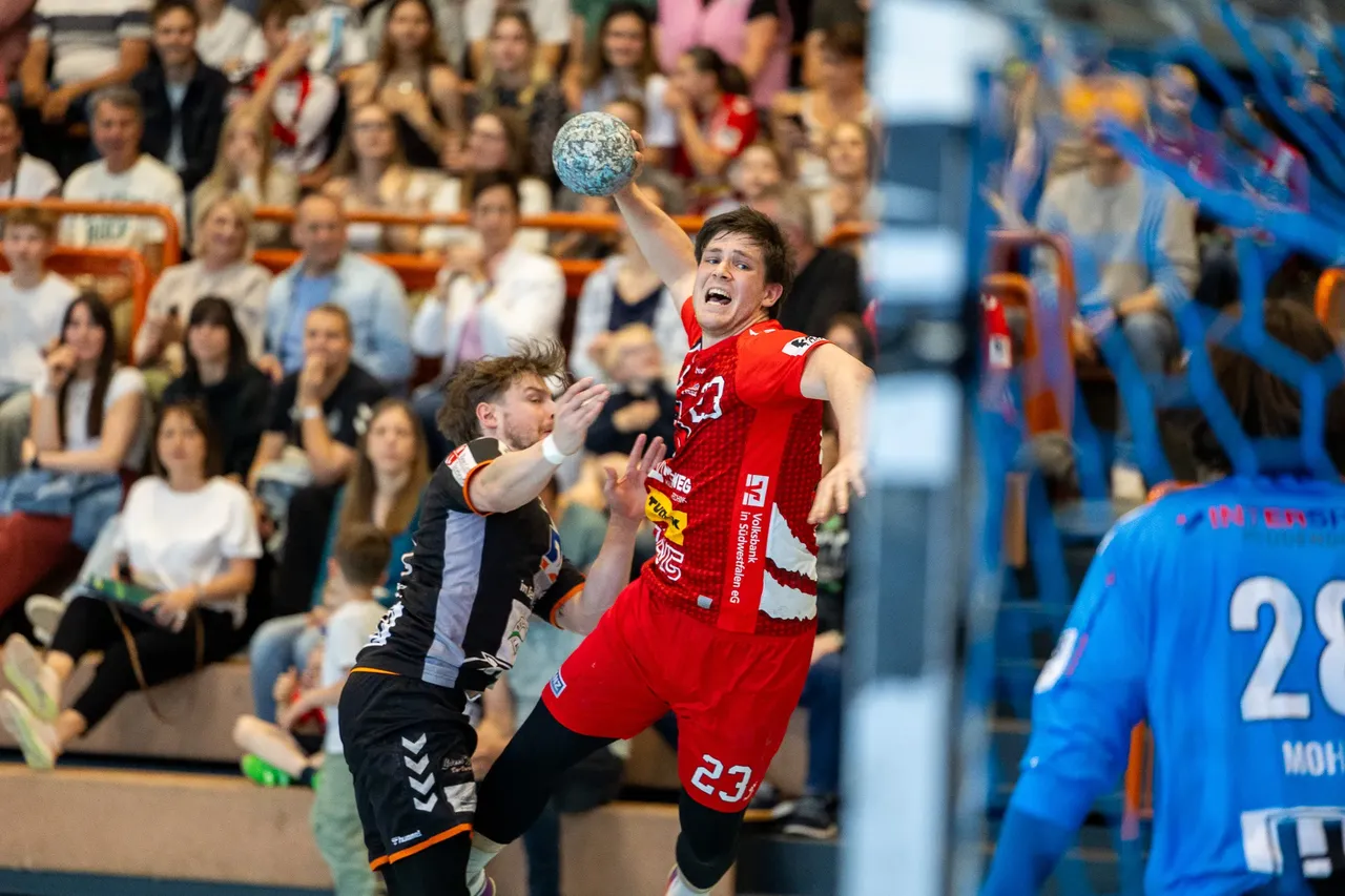 Handballspieler in rotem Trikot wirft den Ball im Spiel gegen die Bergischen Panther, im Hintergrund jubelnde Zuschauer.