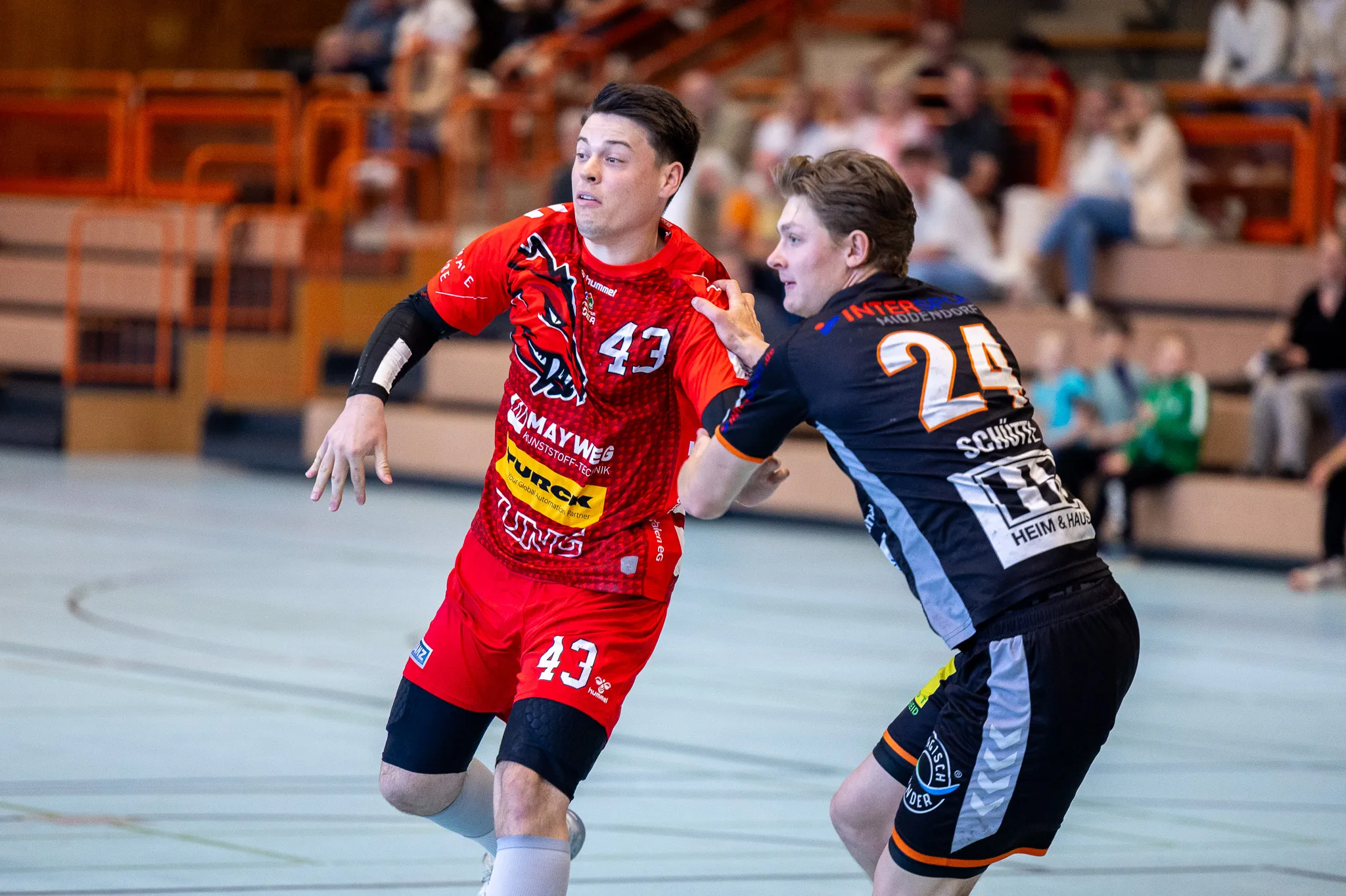 Zwei Handballspieler in Aktion während eines Spiels: Ein Spieler in rotem Trikot mit der Nummer 43 dribbelt den Ball, während ein Gegenspieler in schwarzem Trikot mit der Nummer 24 ihn verteidigt.