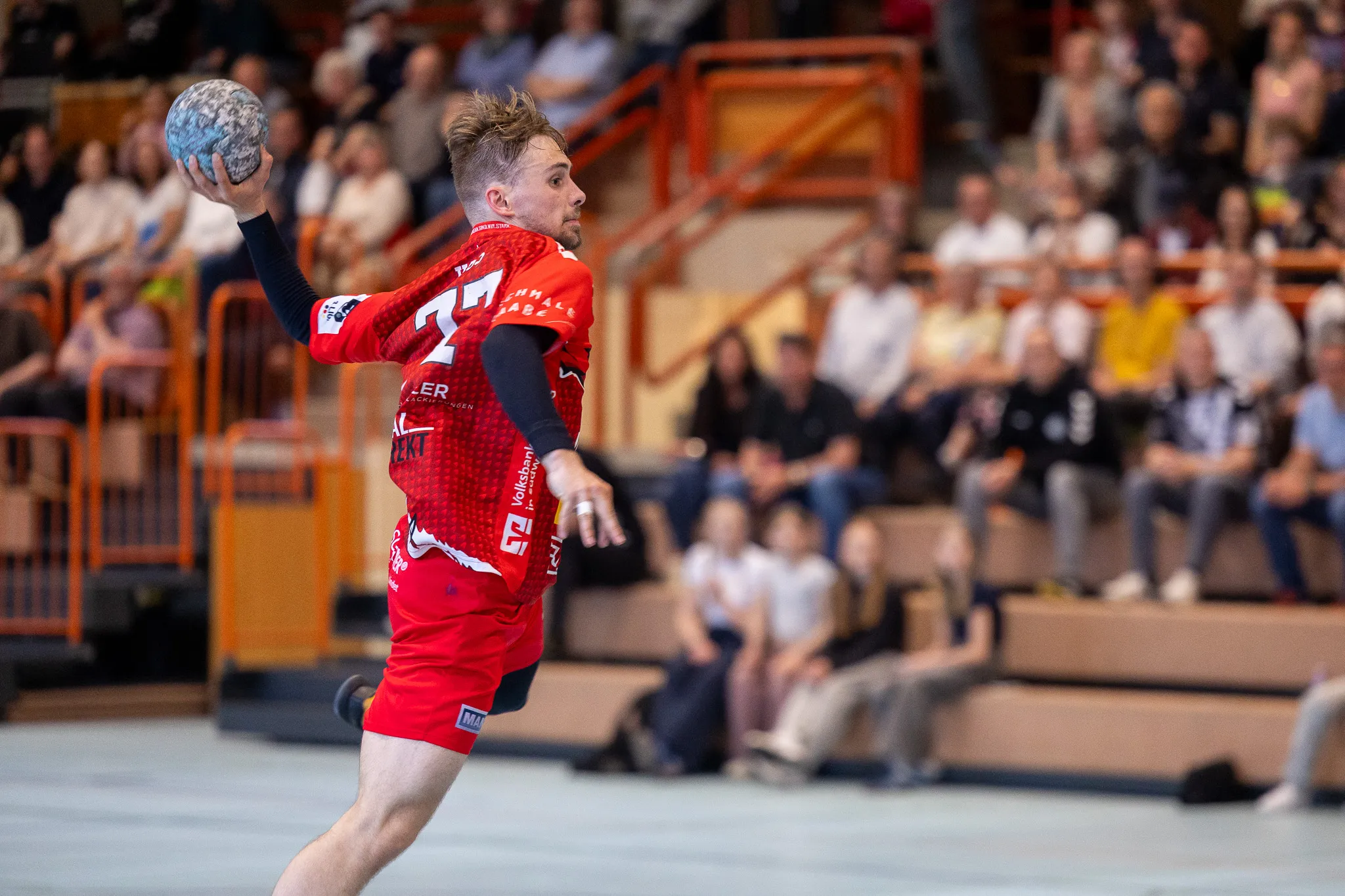 Handballspieler in rotem Trikot wirft den Ball während eines Spiels in einer Halle. Im Hintergrund sind Zuschauer und weitere Spieler zu sehen.