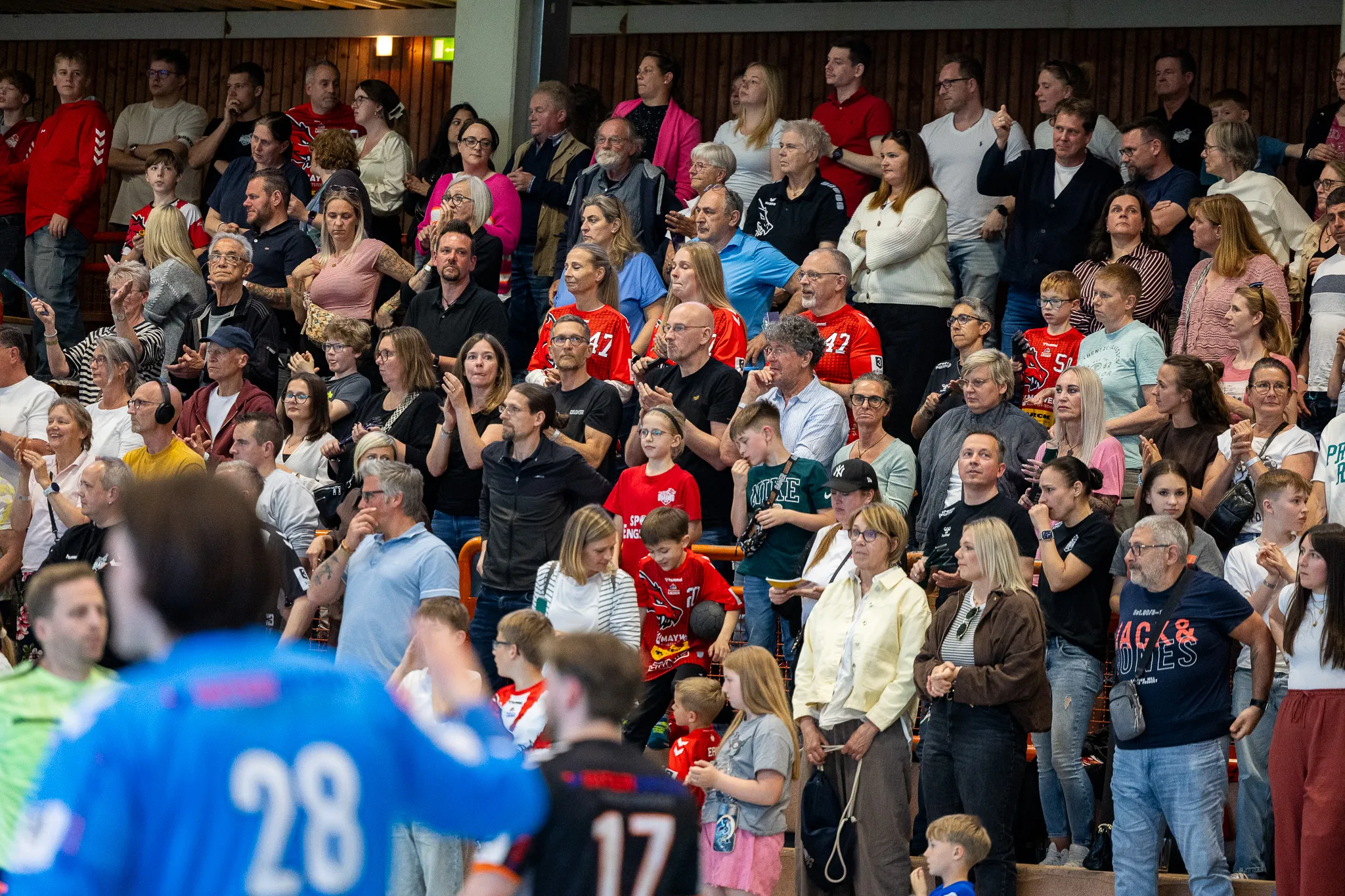 Zuschauer jubeln bei einem Handballspiel in einer Halle, viele tragen rote Trikots mit der Nummer 42.