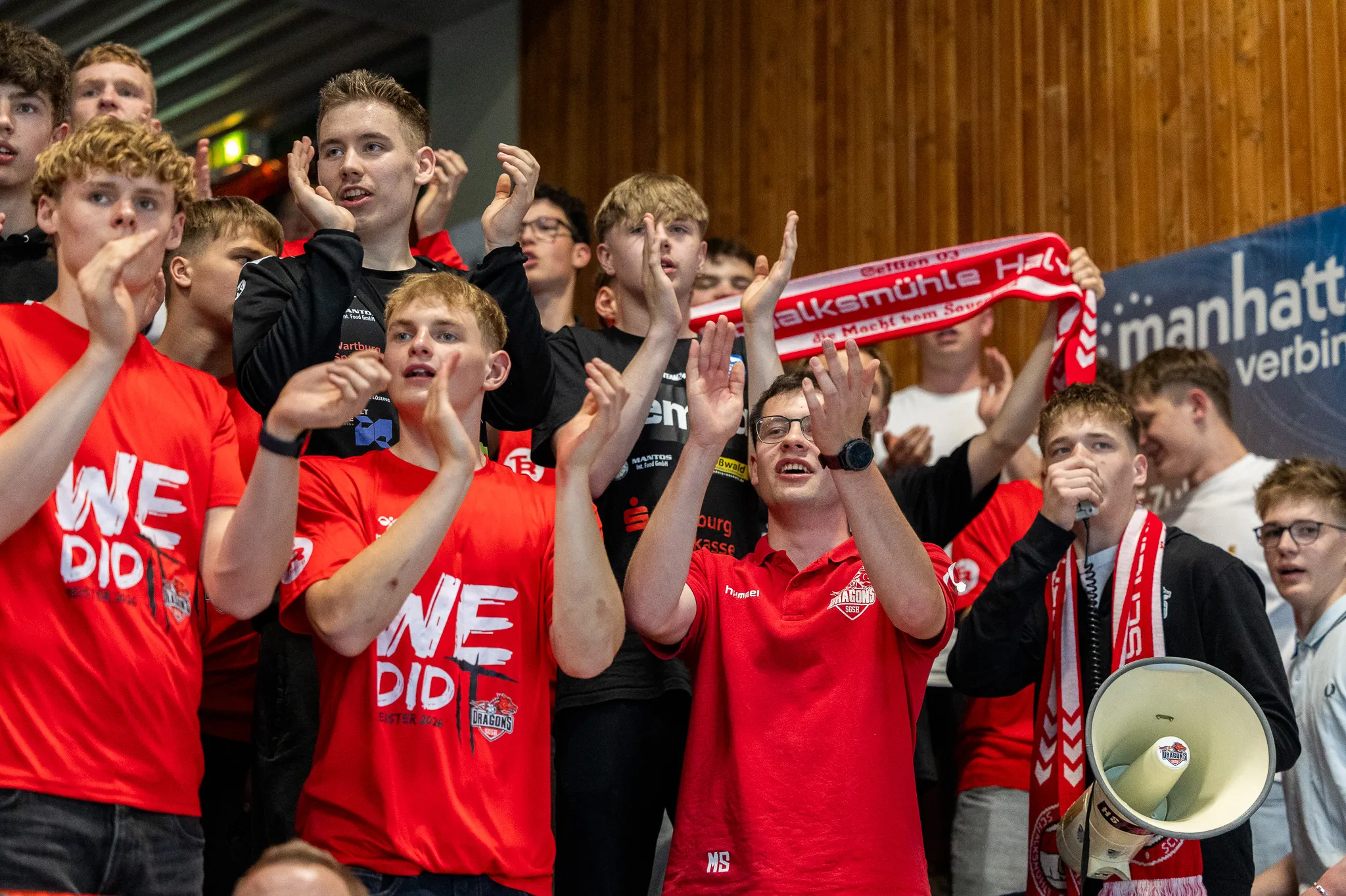 Junge Fans der SGSH Dragons jubeln mit hochgereckten Händen und Schal in den Farben Rot und Weiß. Einige tragen T-Shirts mit dem Aufdruck „WE DID IT“.