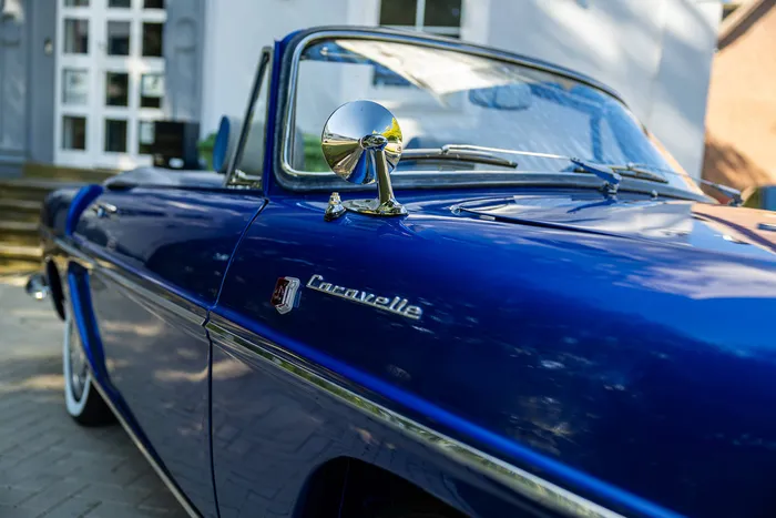 Nahaufnahme eines blauen Oldtimer-Cabriolets mit dem Schriftzug „Caravelle“ und einem verchromten Seitenspiegel.