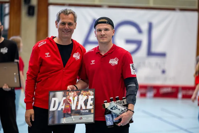 Zwei Männer in roten Trainingsanzügen mit dem Aufdruck „Dragons“ posieren mit einer Collage und einer Auswahl an Bierflaschen.