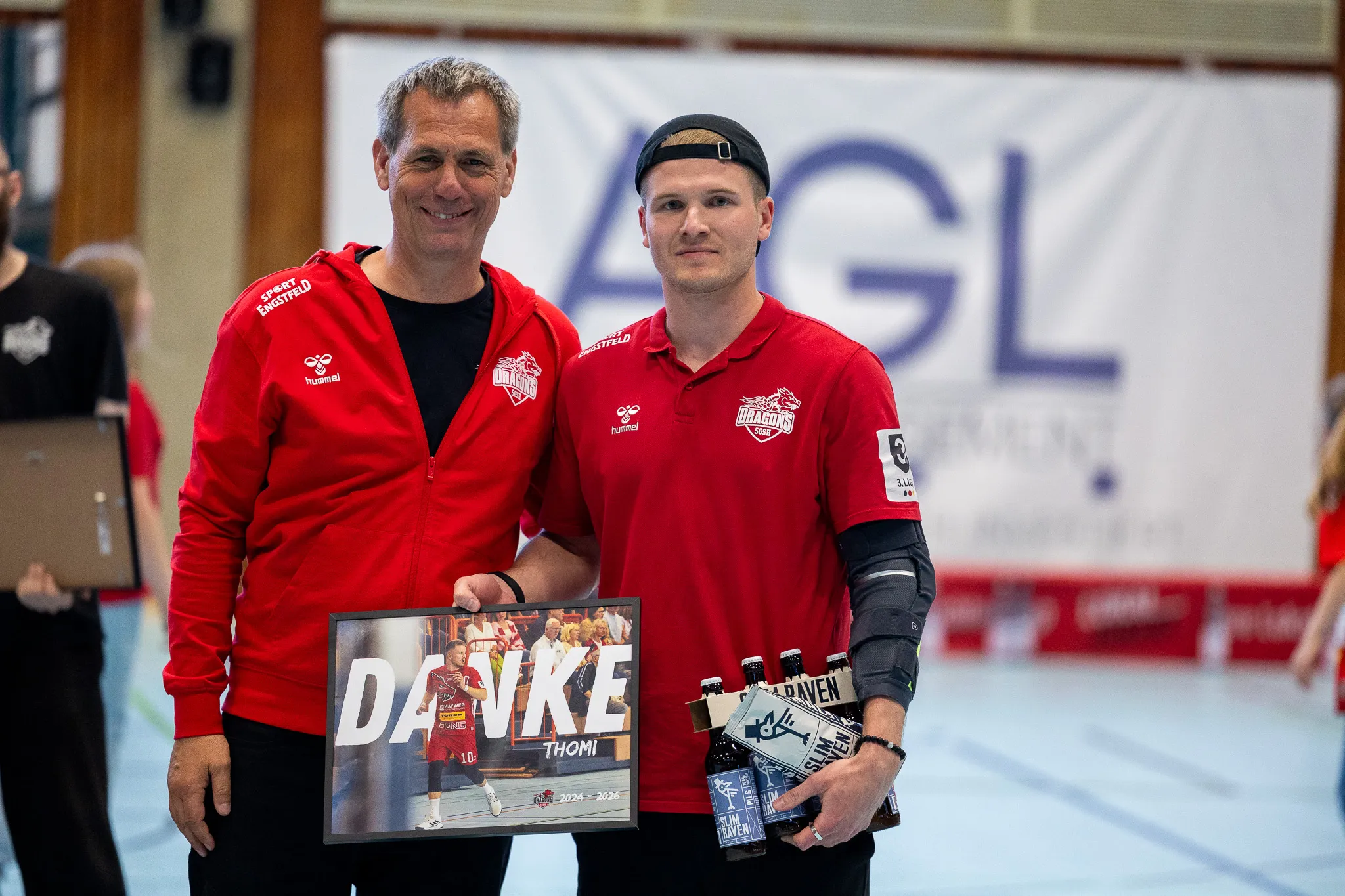 Zwei Männer in roten Trainingsanzügen mit dem Aufdruck „Dragons“ posieren mit einer Collage und einer Auswahl an Bierflaschen.
