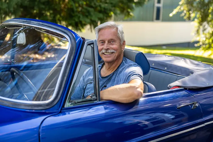 Wilfried Gerdel sitzt lächelnd im Fahrersitz eines blauen Oldtimer-Cabriolets.