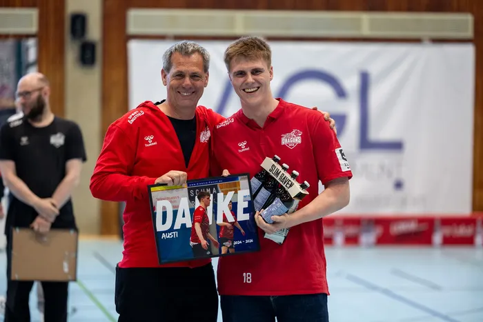 Zwei Männer in roten Trainingsanzügen mit dem Logo „Dragons“ halten Geschenke und eine Dankeskarte mit dem Namen „Austi“.