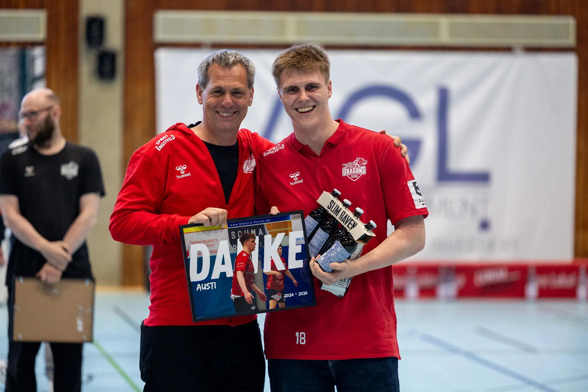 Zwei Männer in roten Trainingsanzügen mit dem Logo „Dragons“ halten Geschenke und eine Dankeskarte mit dem Namen „Austi“.