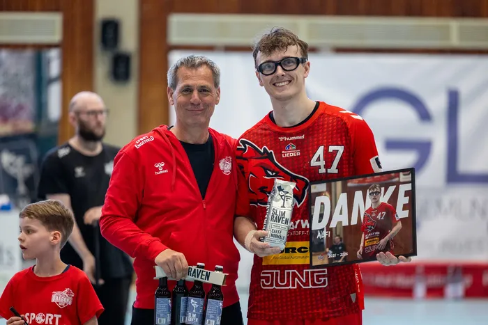 Ein Mann übergibt einem jungen Sportler ein Schild mit der Aufschrift „DANKE“ und dem Logo von Sponsoren in einer Sporthalle.