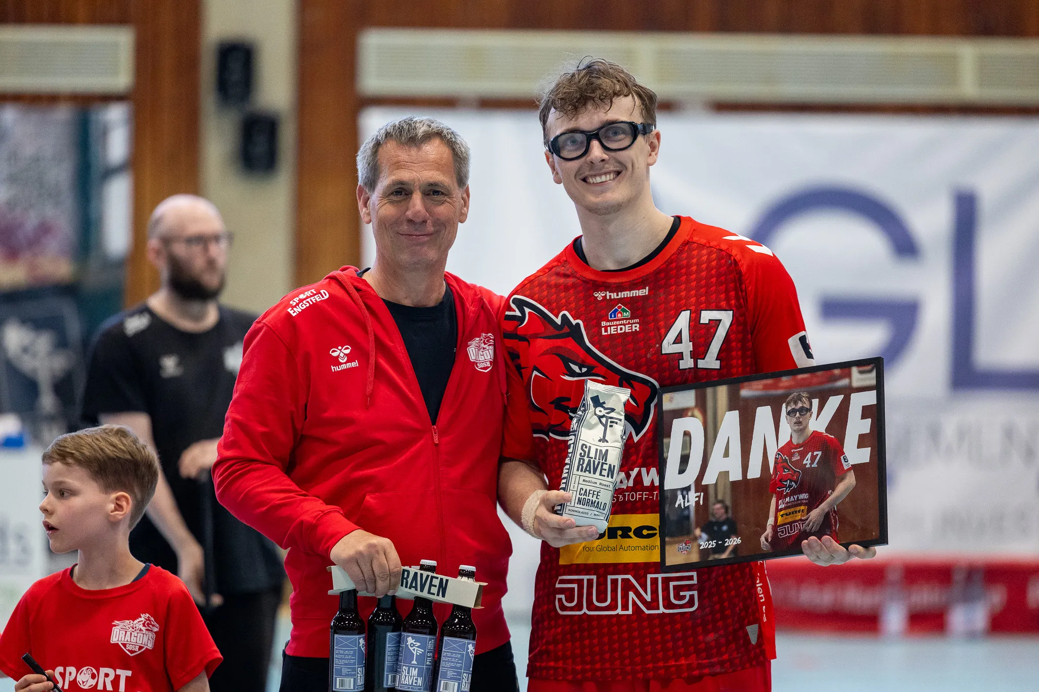 Ein Mann übergibt einem jungen Sportler ein Schild mit der Aufschrift „DANKE“ und dem Logo von Sponsoren in einer Sporthalle.