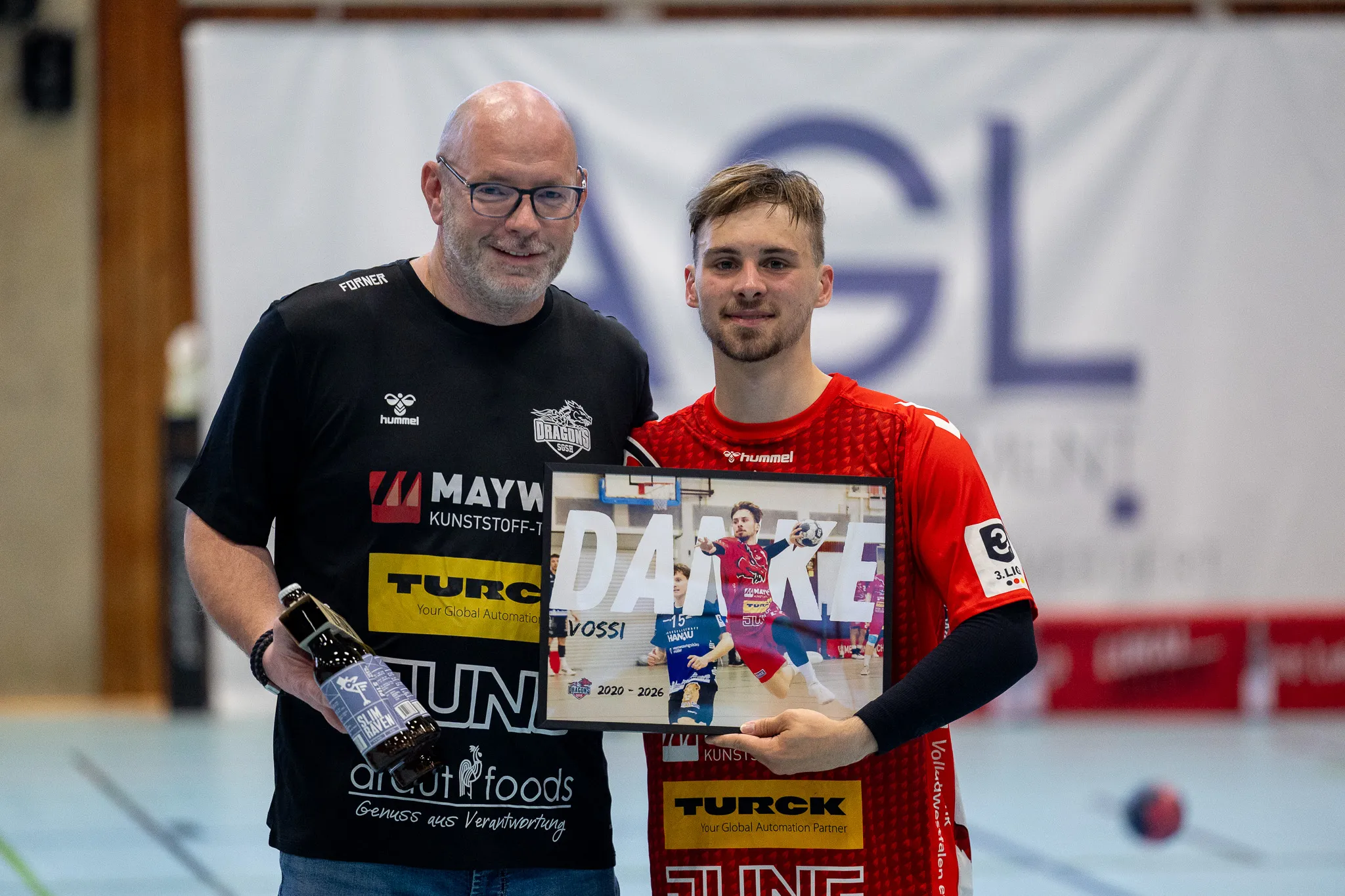 Zwei Männer, ein älterer mit Brille und ein jüngerer, halten eine große Dankestafel mit Fotos und Logos von Sponsoren. Im Hintergrund eine Handballhalle.