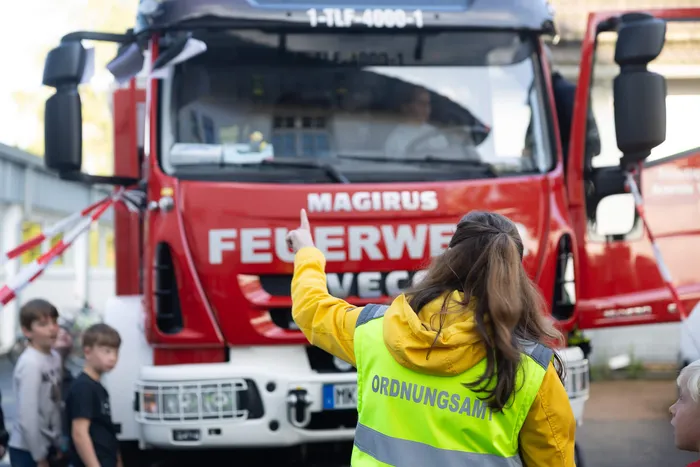Eine Person in einer gelben Warnweste des Ordnungsamtes zeigt auf einen roten Feuerwehrwagen, während Kinder zusehen.