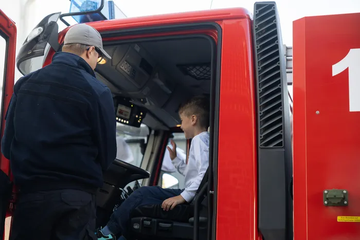 Ein Junge sitzt auf dem Fahrersitz eines roten Feuerwehrautos, ein Mann steht daneben an der offenen Tür.