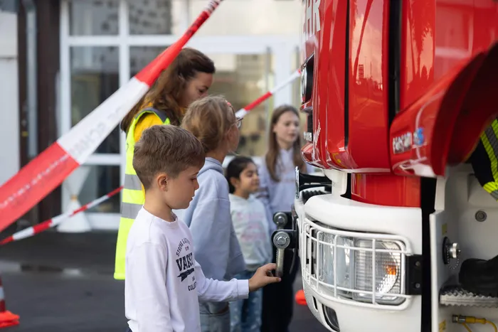 Ein Junge betrachtet den Bereich eines Spiegels an einem roten LKW, während andere Kinder und eine Frau in einer Warnweste zusehen.