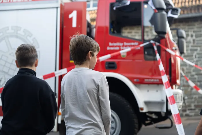 Zwei Jungen stehen mit dem Rücken zur Kamera und betrachten einen roten Feuerwehrwagen hinter rot-weißem Absperrband.