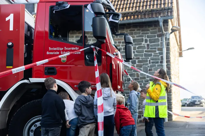 Kinder stehen bei einer Übung zum toten Winkel neben einem roten Feuerwehrwagen der Gemeinde Schalksmühle, der mit Absperrband markiert ist.