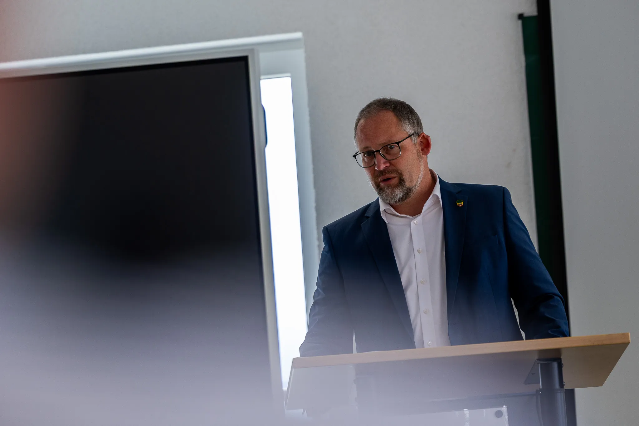 Bürgermeister Armin Kibbert spricht vor einem Publikum, steht an einem Rednerpult und trägt einen dunklen Anzug mit einer Anstecknadel.