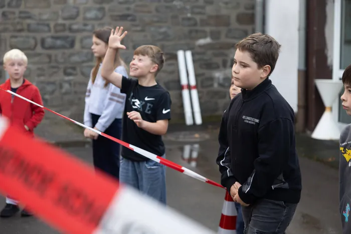 Kinder stehen hinter einem rot-weißen Absperrband bei einer Übung zum Thema „Toter Winkel“, ein Junge hebt die Hand zum Winken.
