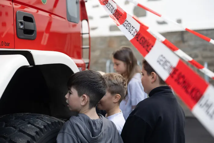 Kinder stehen hinter einem rot-weißen Absperrband und betrachten ein großes rotes Fahrzeug.
