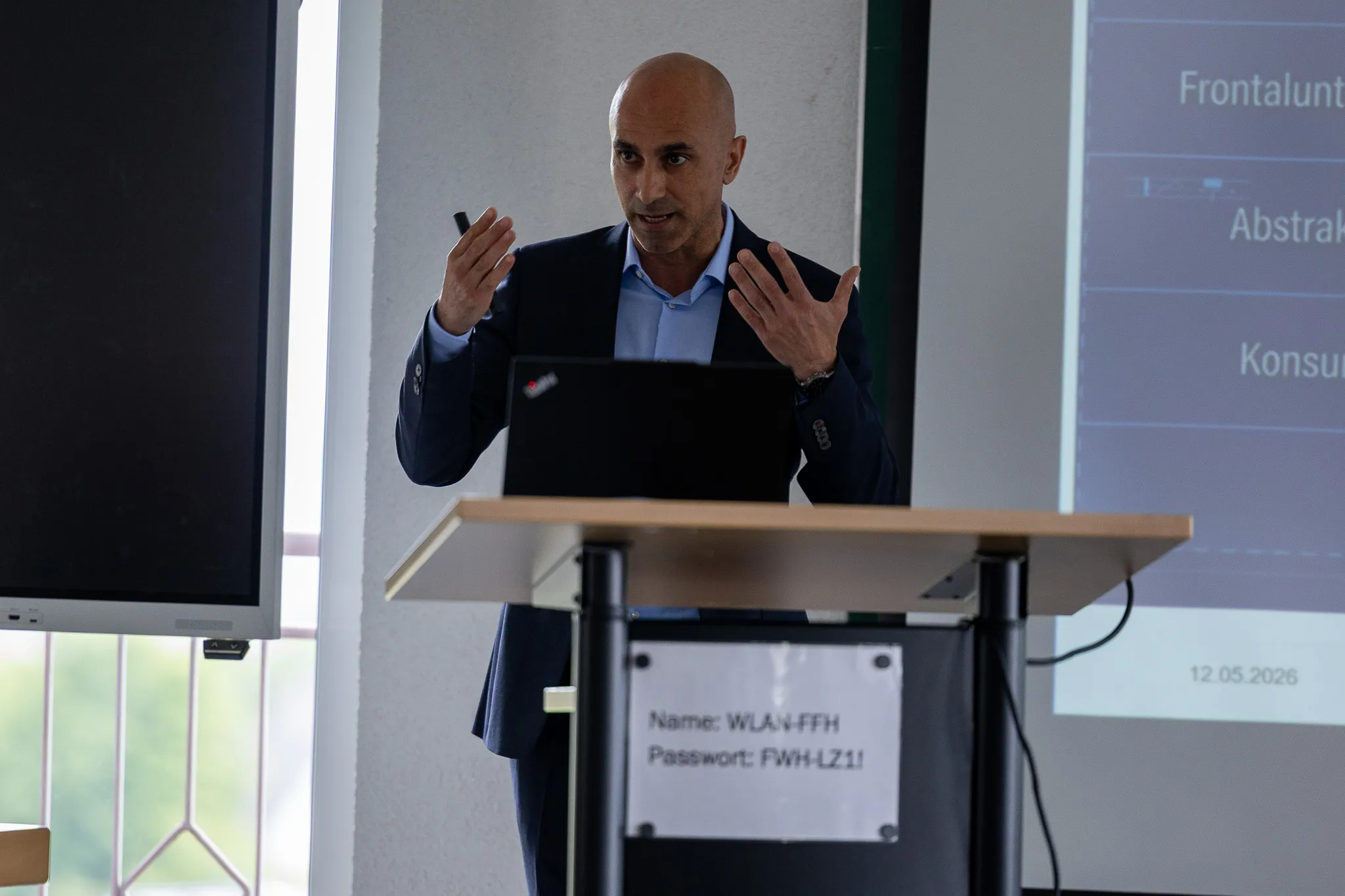 Mann mit Glatze im Anzug gestikuliert während einer Präsentation an einem Rednerpult mit Laptop und WLAN-Informationen.